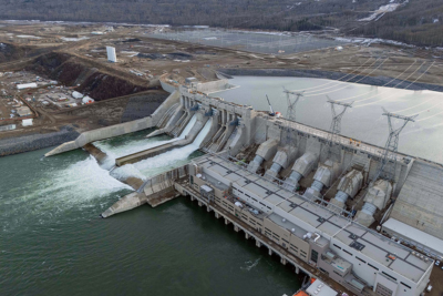 Aerial view of a large hydroelectric dam with multiple spillways releasing water, power lines extending from the facility, and surrounding construction and forested landscape.